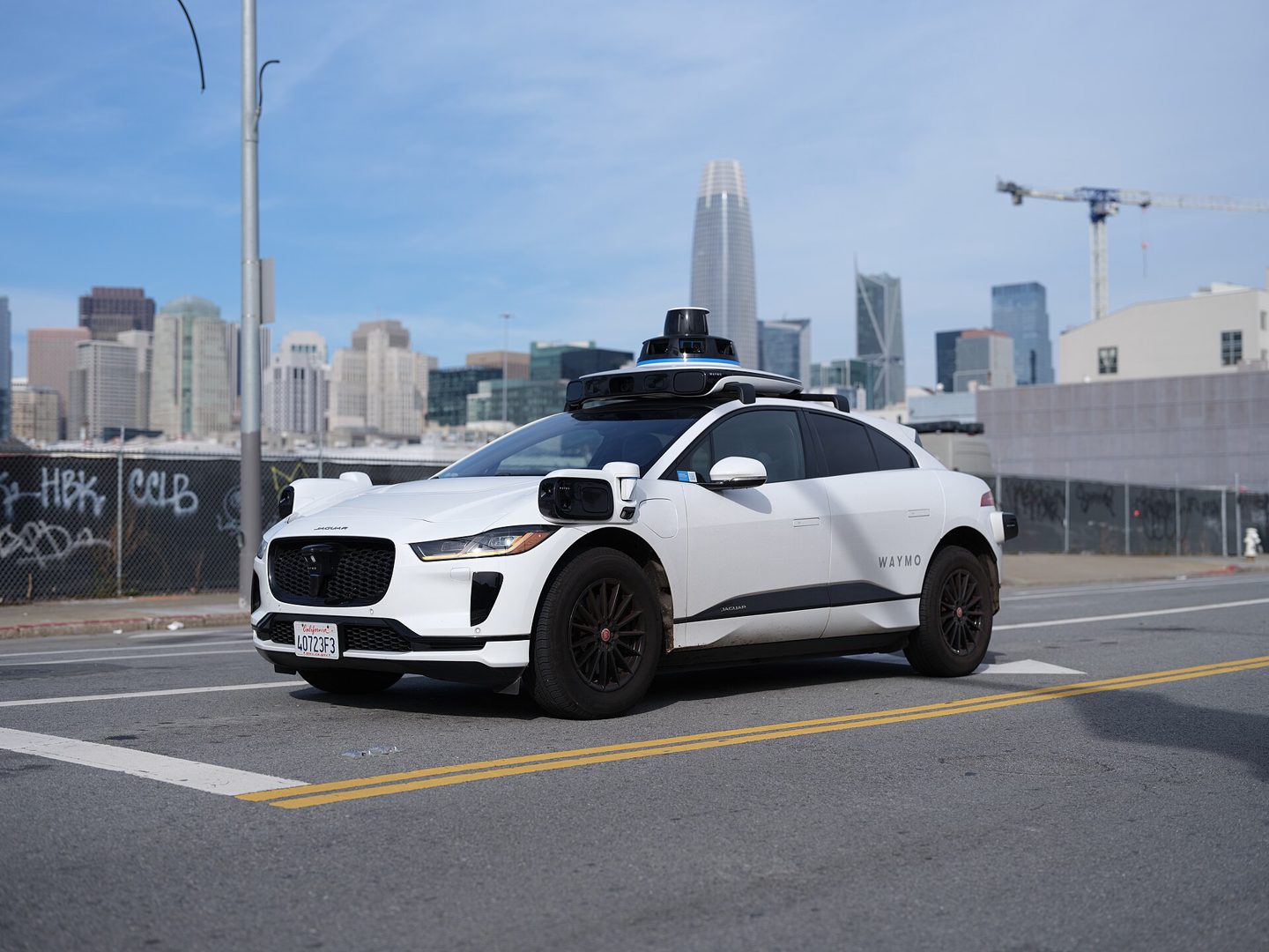 A white Waymo self-driving Jaguar I-PACE with a rooftop lidar sensor array drives through San Francisco with the city skyline behind it.